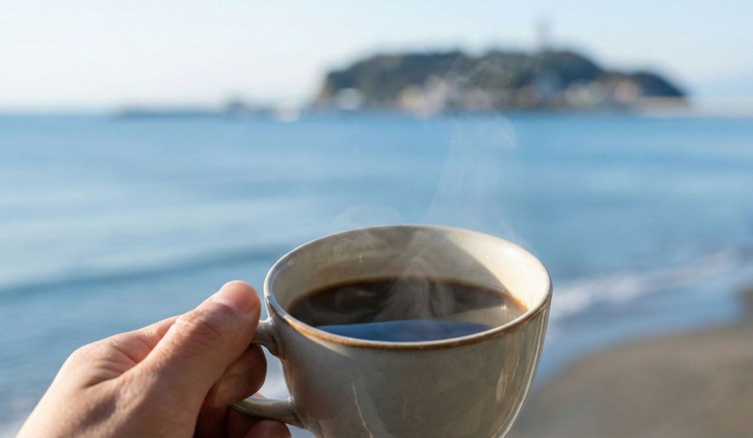 江の島の海を背景に、湯気が立つ温かいコーヒーカップを持つ手元