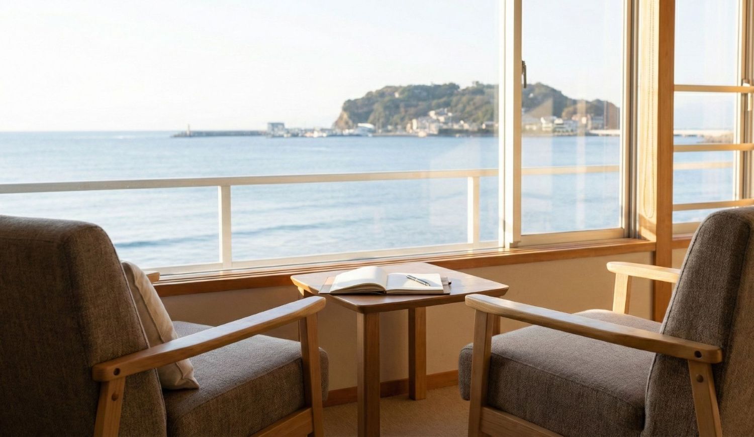 江の島の海が見える窓辺に置かれた二つの椅子と、ノートが開かれたテーブルのある静かな部屋