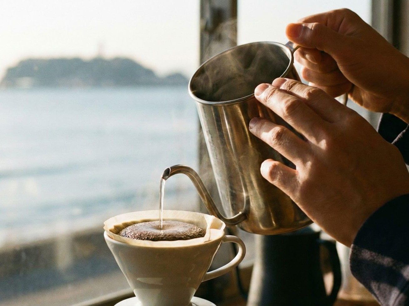 窓の外に広がる江の島の海を背景に、穏やかな光の中で丁寧にコーヒーをハンドドリップする手元のアップ